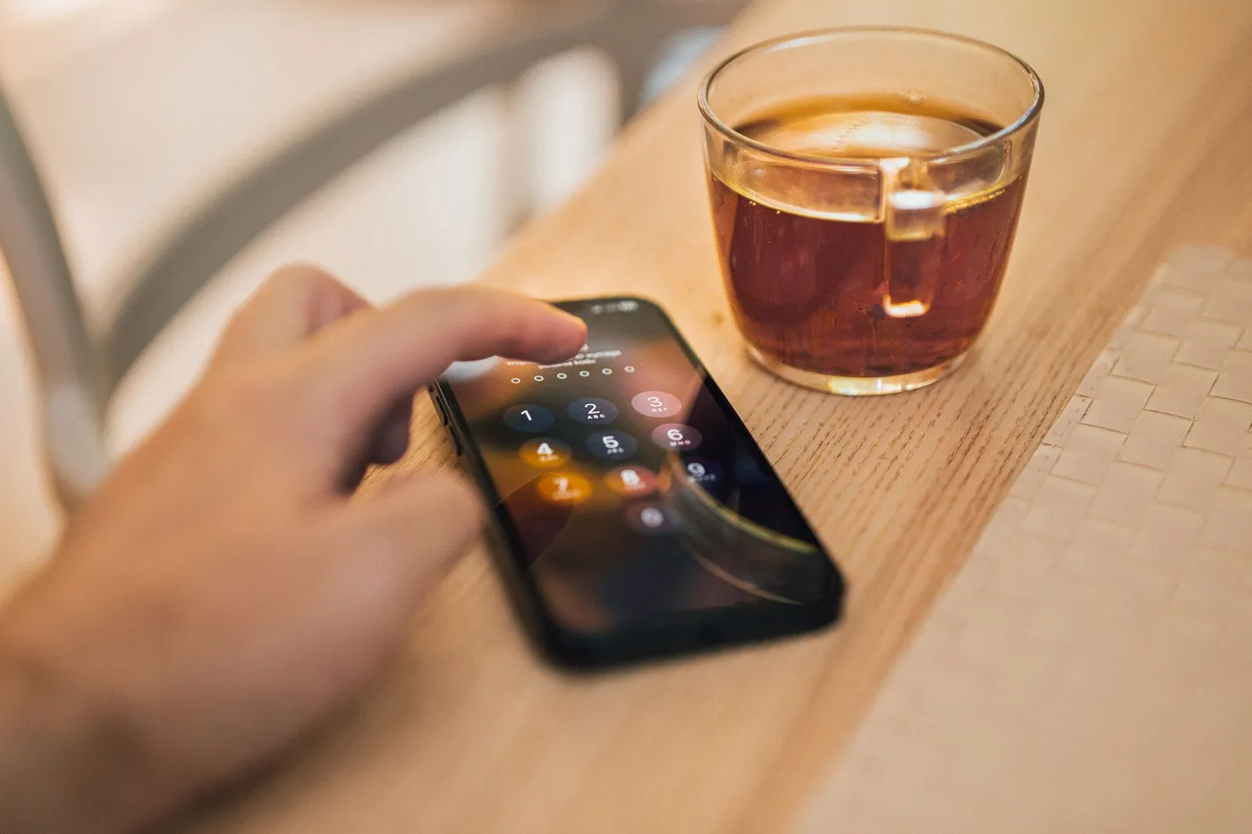 Hand entering a passcode on a smartphone lying on a wooden counter next to a glass of tea