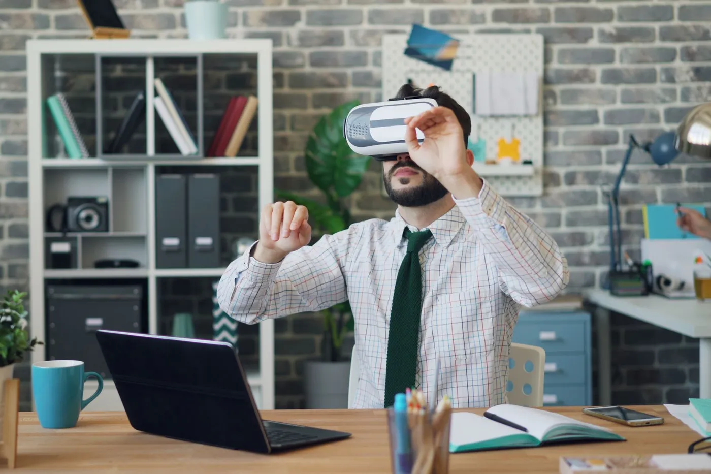 Man at an office desk wearing a white VR headset and reaching out with both hands, an open laptop in front of him