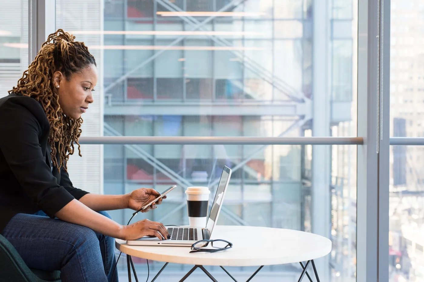 Woman at a round table by a large window typing on a laptop while holding a smartphone